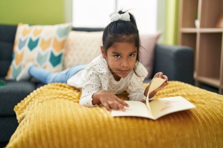 Photo for Adorable hispanic girl reading book lying on sofa at home - Royalty Free Image