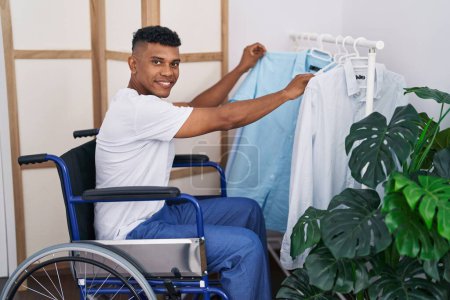 Foto de Hombre latino joven eligiendo camisa sentada en silla de ruedas en el dormitorio - Imagen libre de derechos