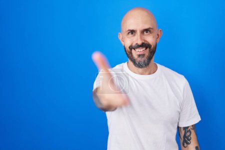 Foto de Hombre hispano con tatuajes de pie sobre fondo azul sonriendo amistoso ofreciendo apretón de manos como saludo y bienvenida. negocios exitosos. - Imagen libre de derechos
