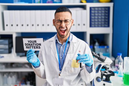 Téléchargez les photos : Young hispanic man working at scientist laboratory holding your donation matters holding blood sample angry and mad screaming frustrated and furious, shouting with anger. rage and aggressive concept. - en image libre de droit