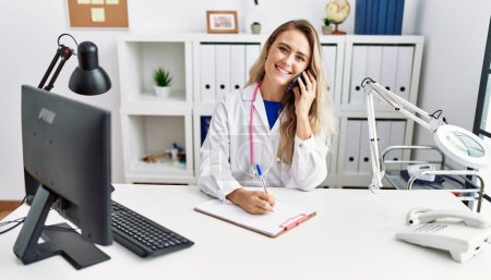 Photo for Young woman wearing doctor uniform writing on document talking on smartphone at clinic - Royalty Free Image