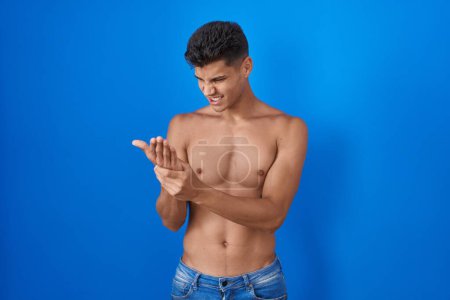 Foto de Joven hombre hispano de pie sin camisa sobre fondo azul sufriendo dolor en manos y dedos, inflamación de la artritis - Imagen libre de derechos