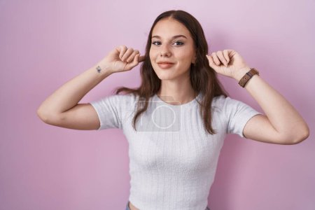 Téléchargez les photos : Jeune fille hispanique debout sur fond rose souriant tirant les oreilles avec les doigts, geste drôle. problème d'audition - en image libre de droit