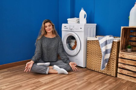 Photo for Young blonde woman doing yoga exercise waiting for washing machine at laundry room - Royalty Free Image