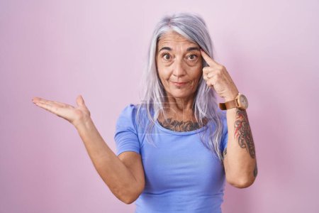 Photo for Middle age woman with tattoos standing over pink background confused and annoyed with open palm showing copy space and pointing finger to forehead. think about it. - Royalty Free Image