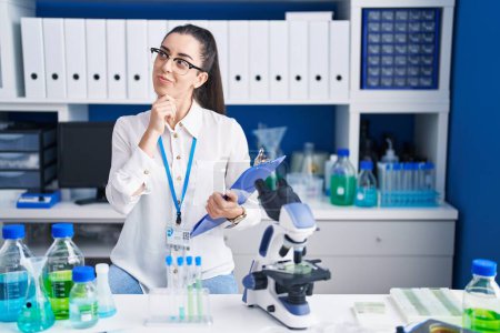 Photo for Young brunette woman working at scientist laboratory with hand on chin thinking about question, pensive expression. smiling with thoughtful face. doubt concept. - Royalty Free Image