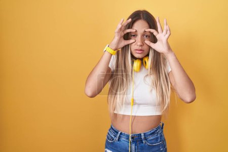 Téléchargez les photos : Young blonde woman standing over yellow background wearing headphones trying to open eyes with fingers, sleepy and tired for morning fatigue - en image libre de droit