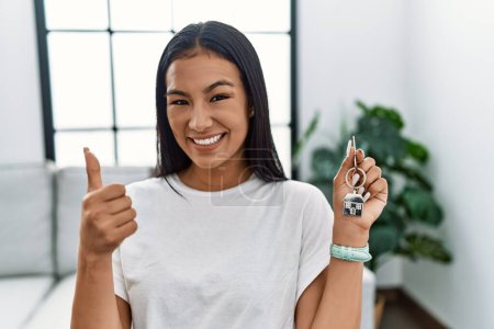 Foto de Young hispanic woman holding keys of new home doing happy thumbs up gesture with hand. approving expression looking at the camera showing success. - Imagen libre de derechos