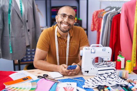 Téléchargez les photos : Jeune homme latino tailleur souriant confiant en utilisant smartphone à l'atelier - en image libre de droit