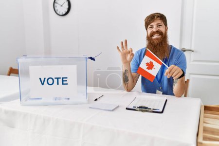 Foto de Hombre caucásico con barba larga en las elecciones de campaña política sosteniendo la bandera de Canadá haciendo señal bien con los dedos, sonriendo gesto amistoso excelente símbolo - Imagen libre de derechos