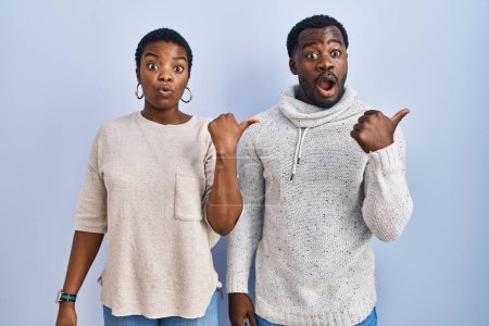 Photo for Young african american couple standing over blue background together surprised pointing with hand finger to the side, open mouth amazed expression. - Royalty Free Image