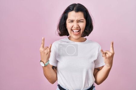 Téléchargez les photos : Young hispanic woman wearing casual white t shirt over pink background shouting with crazy expression doing rock symbol with hands up. music star. heavy music concept. - en image libre de droit