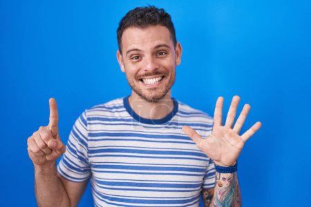 Foto de Young hispanic man standing over blue background showing and pointing up with fingers number six while smiling confident and happy. - Imagen libre de derechos
