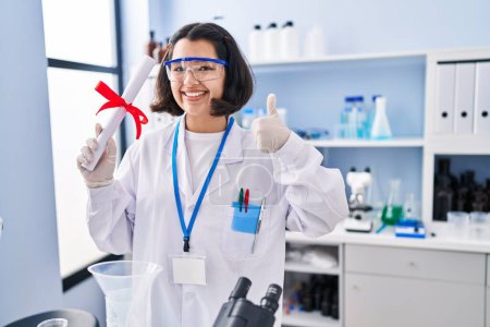 Photo for Young hispanic woman working at scientist laboratory holding degree smiling happy and positive, thumb up doing excellent and approval sign - Royalty Free Image
