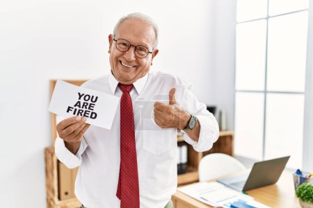 Photo for Senior business man holding you are fired banner at the office smiling happy and positive, thumb up doing excellent and approval sign - Royalty Free Image