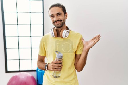 Photo for Young hispanic man wearing sportswear and drinking water at the gym pointing aside with hands open palms showing copy space, presenting advertisement smiling excited happy - Royalty Free Image
