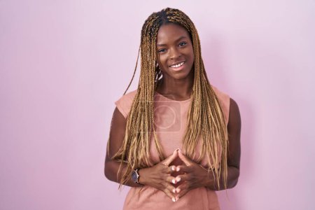 Téléchargez les photos : Femme afro-américaine avec les cheveux tressés debout sur fond rose mains ensemble et doigts croisés souriant détendu et joyeux. succès et optimisme - en image libre de droit