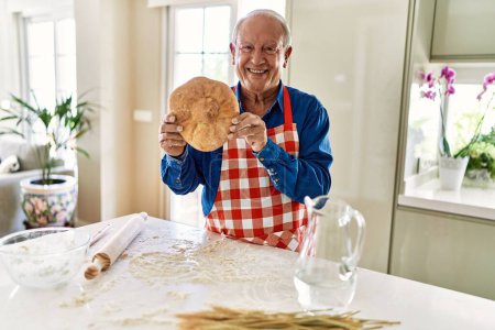 Foto de Hombre mayor sonriendo confiado sosteniendo pan casero en la cocina - Imagen libre de derechos
