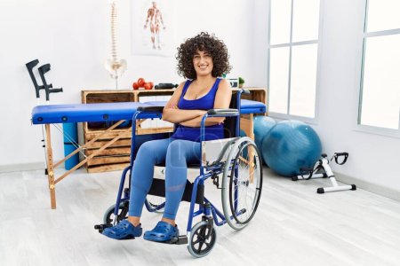 Téléchargez les photos : Young middle eastern woman sitting on wheelchair at physiotherapy clinic happy face smiling with crossed arms looking at the camera. positive person. - en image libre de droit
