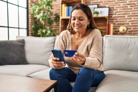 Foto de Mujer hispana de mediana edad usando teléfono inteligente y tarjeta de crédito sentada en el sofá en casa - Imagen libre de derechos
