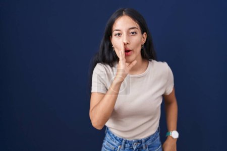Téléchargez les photos : Jeune femme hispanique debout sur fond bleu main sur bouche racontant rumeur secrète, chuchotant conversation malicieuse - en image libre de droit