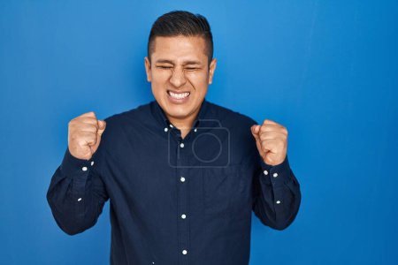 Photo for Hispanic young man standing over blue background excited for success with arms raised and eyes closed celebrating victory smiling. winner concept. - Royalty Free Image