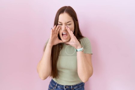 Foto de Hermosa mujer morena de pie sobre fondo rosa gritando enojado en voz alta con las manos sobre la boca - Imagen libre de derechos