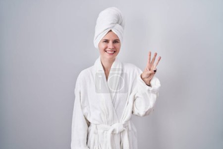 Téléchargez les photos : Blonde femme caucasienne portant peignoir montrant et pointant vers le haut avec les doigts numéro trois tout en souriant confiant et heureux. - en image libre de droit
