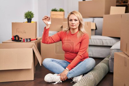 Photo for Blonde woman holding keys of new home sitting on the floor thinking attitude and sober expression looking self confident - Royalty Free Image
