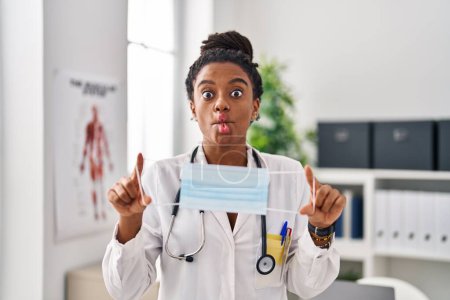 Photo for Young african american with braids wearing doctor uniform holding safety mask making fish face with mouth and squinting eyes, crazy and comical. - Royalty Free Image