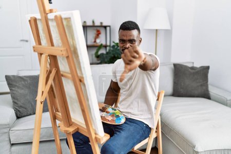 Foto de Young african man painting on canvas at home looking unhappy and angry showing rejection and negative with thumbs down gesture. bad expression. - Imagen libre de derechos