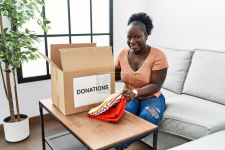 Téléchargez les photos : Jeune femme afro-américaine pliant des vêtements à donner à la maison - en image libre de droit