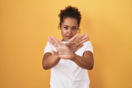 Téléchargez les photos : Young hispanic woman with curly hair standing over yellow background rejection expression crossing arms and palms doing negative sign, angry face - en image libre de droit