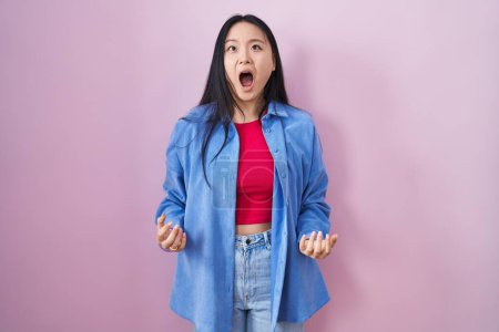 Foto de Mujer asiática joven de pie sobre fondo rosa enojado y loco gritando frustrado y furioso, gritando con ira. rabia y concepto agresivo. - Imagen libre de derechos