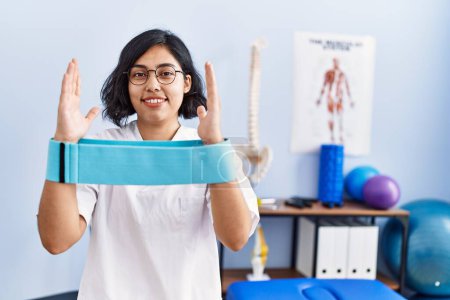 Foto de Young latin woman wearing physiotherapist uniform holding elastic band at physiotherapy clinic - Imagen libre de derechos