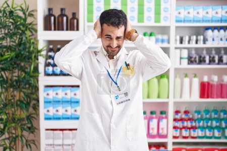 Photo for Handsome hispanic man working at pharmacy drugstore suffering from headache desperate and stressed because pain and migraine. hands on head. - Royalty Free Image