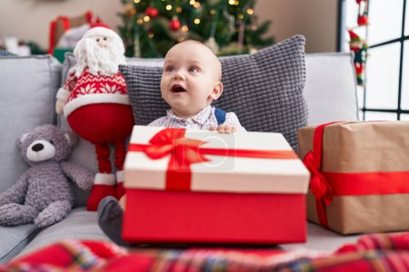 Foto de Adorable bebé caucásico sosteniendo regalo sentado en el sofá por el árbol de Navidad en casa - Imagen libre de derechos