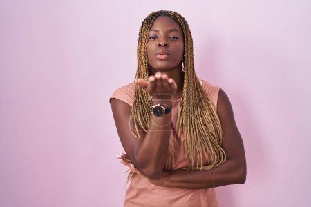 Téléchargez les photos : Femme afro-américaine avec les cheveux tressés debout sur fond rose regardant la caméra souffler un baiser avec la main sur l'air étant belle et sexy. expression d'amour. - en image libre de droit