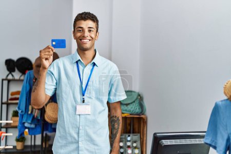 Téléchargez les photos : Jeune homme hispanique travaillant comme assistant de magasin détenant une carte de crédit au magasin de détail - en image libre de droit