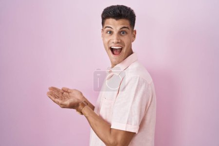 Foto de Joven hombre hispano de pie sobre fondo rosa señalando a un lado con las manos abiertas palmeras mostrando espacio de copia, presentando anuncio sonriente emocionado feliz - Imagen libre de derechos