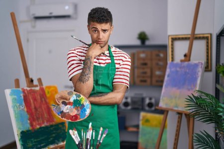 Foto de Joven brasileño sosteniendo paleta de pintores en el estudio de artista escéptico y nervioso, frunciendo el ceño molesto por problemas. persona negativa. - Imagen libre de derechos