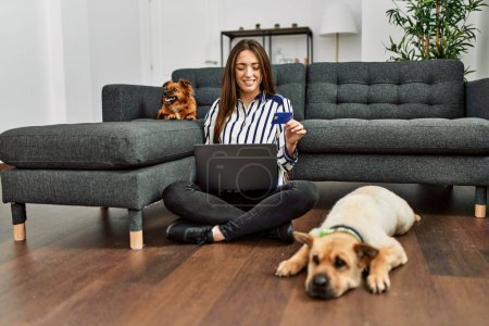 Foto de Joven mujer hispana usando laptop y tarjeta de crédito sentada en el piso con perros en casa - Imagen libre de derechos