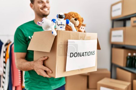 Photo for Young hispanic man wearing volunteer uniform holding box with toys at charity center - Royalty Free Image