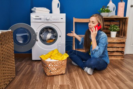 Téléchargez les photos : Jeune femme blonde parlant sur smartphone pour machine à laver cassée à la buanderie - en image libre de droit