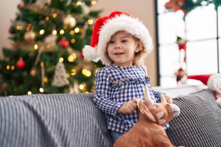 Photo for Adorable hispanic girl holding toy standing on sofa by christmas tree at home - Royalty Free Image