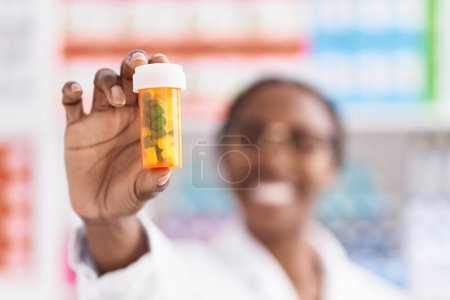 Téléchargez les photos : Afro-américaine femme pharmacien souriant confiant tenant bouteille pilules à la pharmacie - en image libre de droit