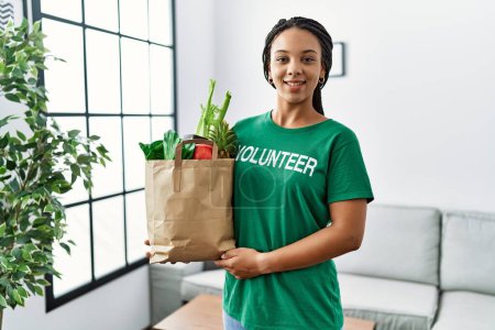 Photo for Young african american woman wearing volunteer t shirt holding bag of groceries looking positive and happy standing and smiling with a confident smile showing teeth - Royalty Free Image