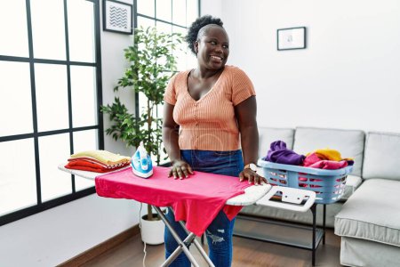 Téléchargez les photos : Jeune femme africaine repassage vêtements à la maison regardant de côté avec le sourire sur le visage, expression naturelle. riant confiant. - en image libre de droit