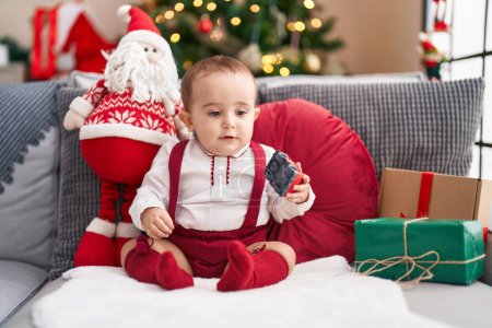 Foto de Adorable bebé hispano sosteniendo juguete de coche sentado en el sofá por el árbol de Navidad en casa - Imagen libre de derechos