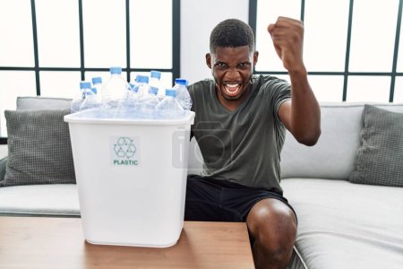 Foto de Joven afroamericano sosteniendo cesto de basura con botellas de plástico de reciclaje molesto y frustrado gritando con ira, gritando loco de ira y levantado a mano - Imagen libre de derechos